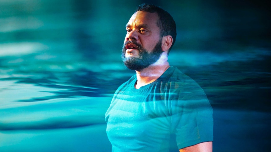 An Aboriginal man with a trim black beard. He's wearing a blue t-shirt and standing in front of a projection of water.