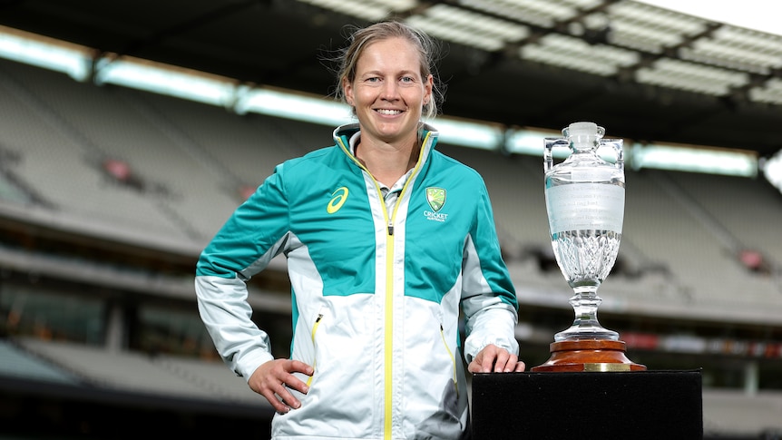 A smiling cricketer has her left hand on a plinth holding the Women' Ashes trophy.