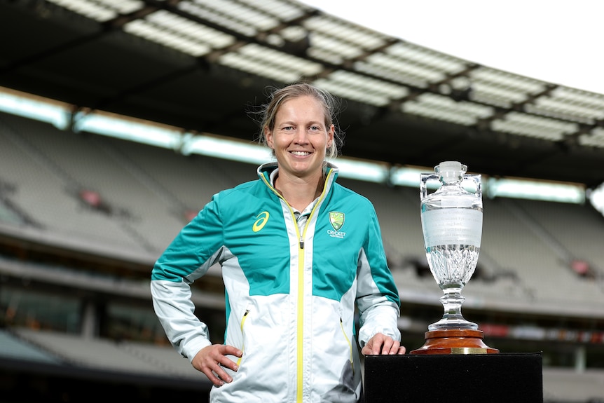 A smiling cricketer has her left hand on a plinth holding the Women' Ashes trophy.