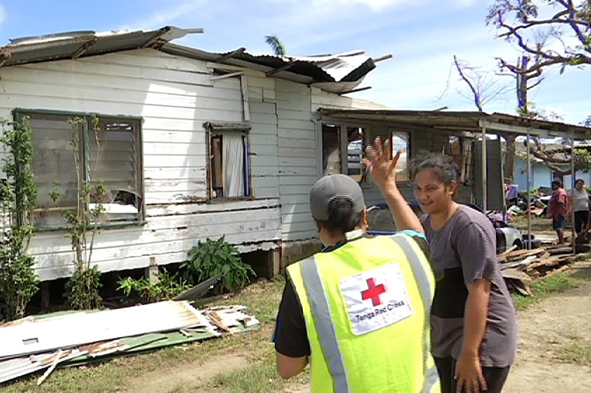 A Red Cross volunteer speaks to a woman with a damaged house in the background.