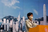 A middle-aged woman wearing a mask and business suit stands at podium with city skyline behind her.