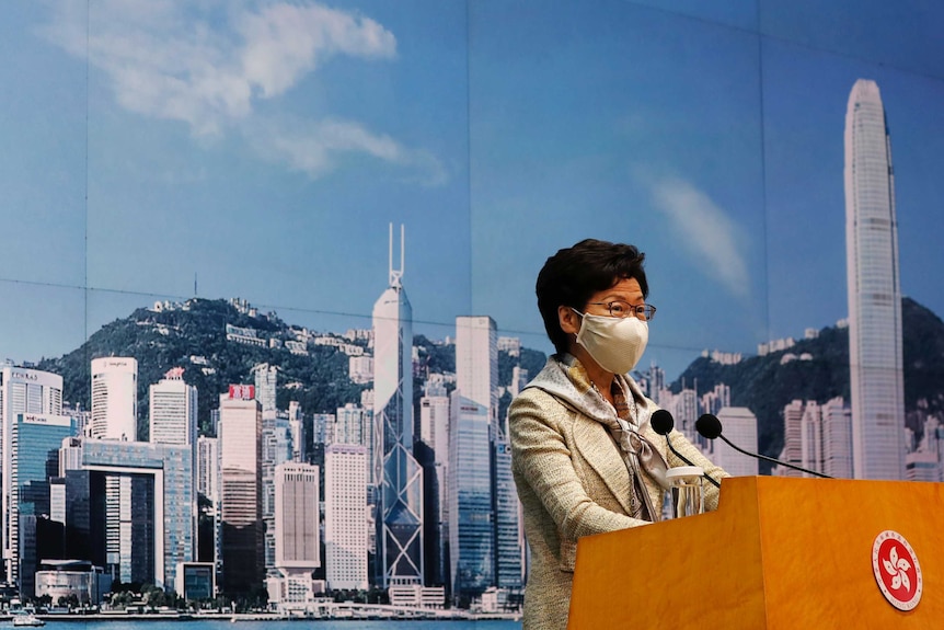 A middle-aged woman wearing a mask and business suit stands at podium with city skyline behind her.