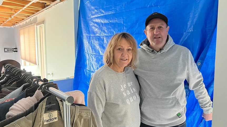 couple in grey jumpers stand together inside storm damaged home