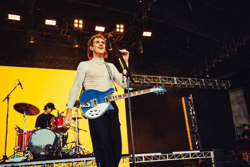 A man with a guitar stands behind a microphone on a stage with a drummer in the background