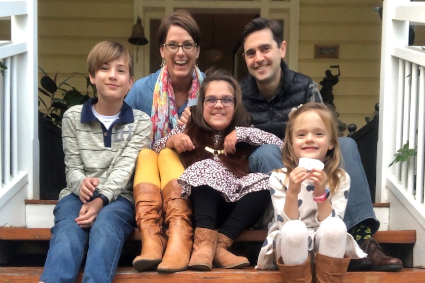 A mum and dad with three young children sit on the front stairs of their home.