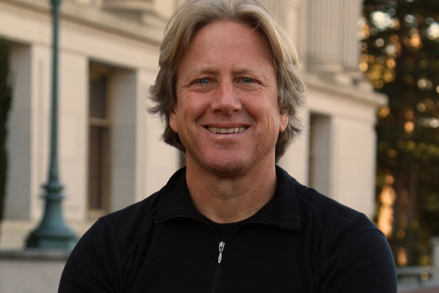 Dacher Keltner, who has blonde-brown hair, poses for a photo in a dark pullover, with his arms folded, smiling