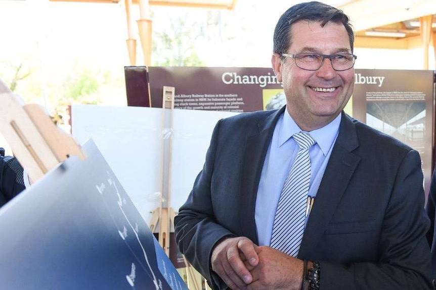 A man with glasses and dressed in a suit stands smiling with signs about Albury behind and next to him.