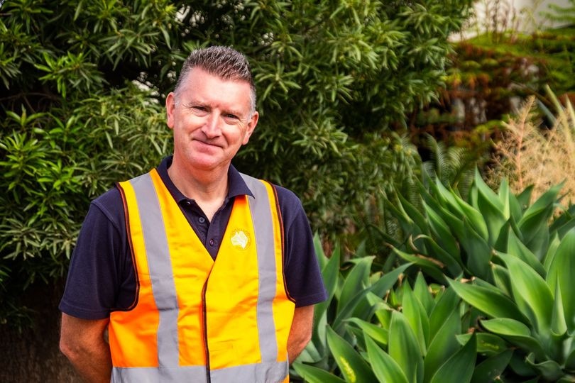A man with short hair wearing a navy polo and orange hi-vis vest stands in front of green plants.