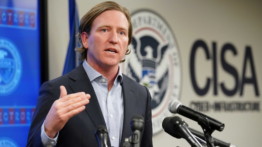 A man wearing a suit without a tie gestures with his right hand as he speaks from behind a lectern in a briefing room.