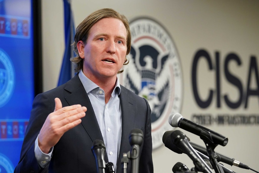 A man wearing a suit without a tie gestures with his right hand as he speaks from behind a lectern in a briefing room.