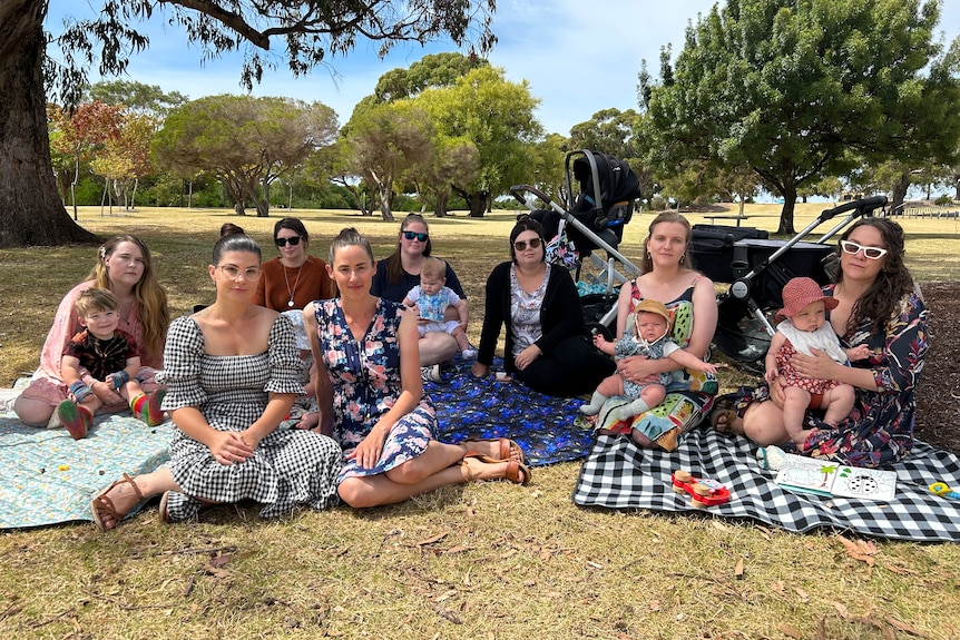 A group of women and babies sit on picnic mats on grass under tress.
