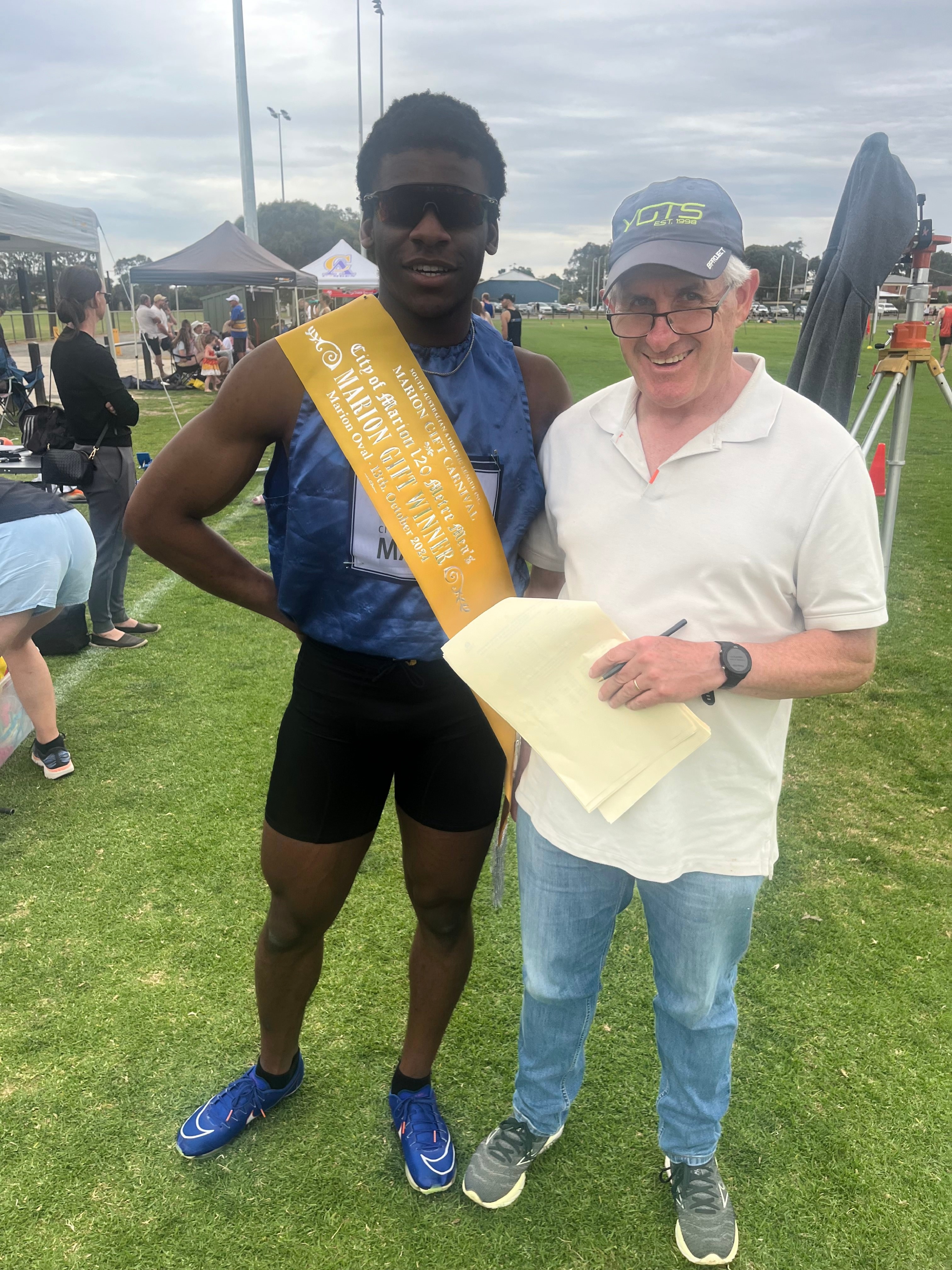 A man in a blue singlet and wearing a winner sash standing next to an older man in a white shirt, glasses and a cap.