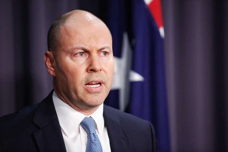 Josh Frydenberg in a suit and tie standing in front of a lecturn in a blue room in front of an australian flag