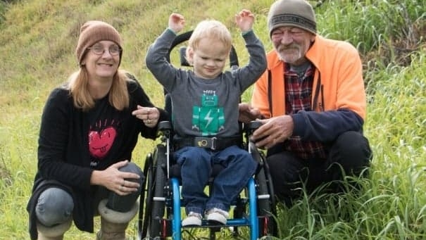 A woman and a man sit smiling in the grass with their young son has special needs in a wheelchair