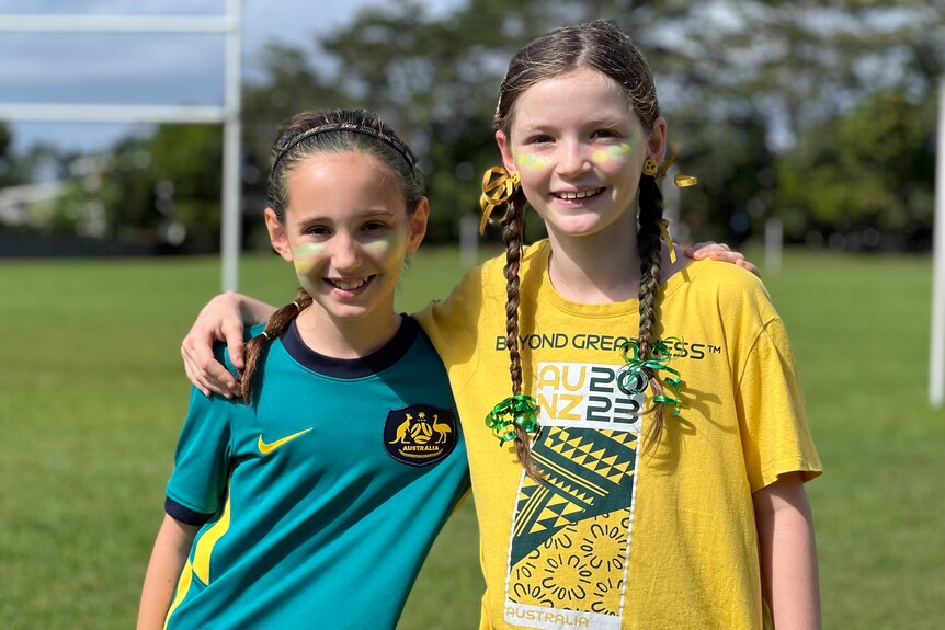 Two girls wearing green and gold supporter gear standing on a football field