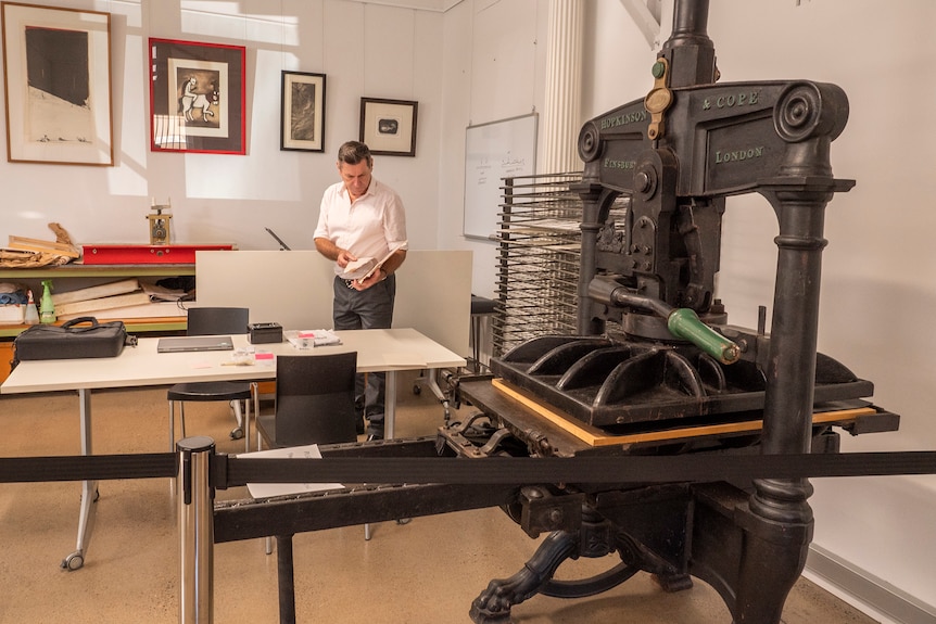 An antique printing press in the foreground with a man standing behind it.