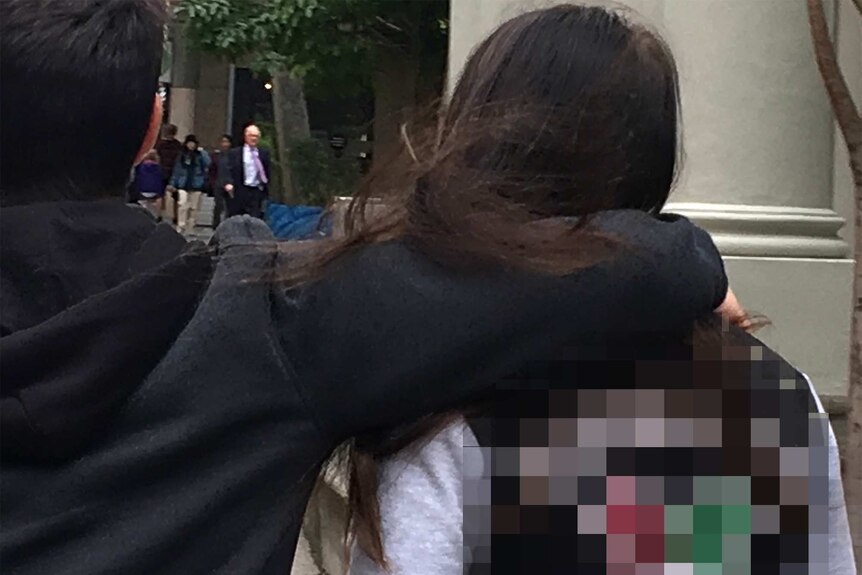 A woman walks down a street photographed from behind with a man walking next to her with his arm on her shoulders.