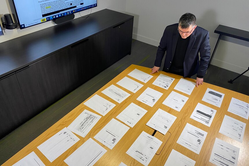 A man in a dark jacket stands in front of reams of paper spread out on a boardroom table.
