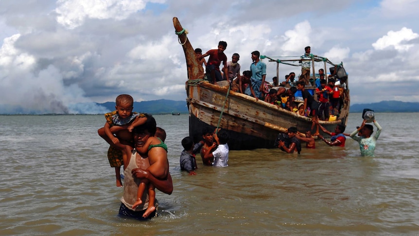 A man carries two children on his shoulders through water, while behind him is a boat carrying others