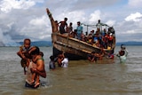 A man carries two children on his shoulders through water, while behind him is a boat carrying others