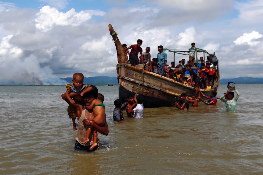 A man carries two children on his shoulders through water, while behind him is a boat carrying others