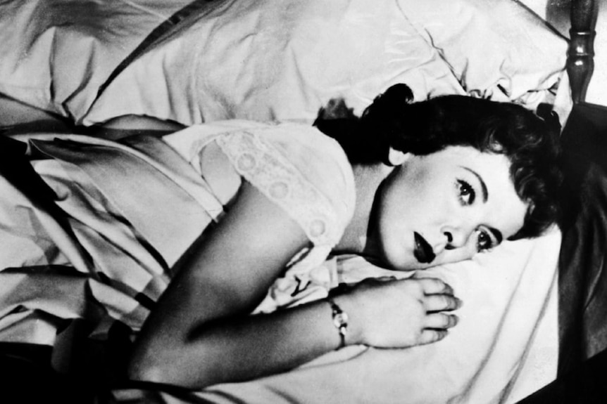 A woman with 1950s style haircut and make-up lies stomach down on bed wide awake.