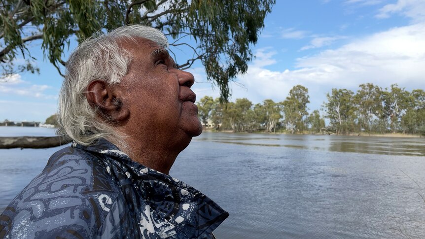 An Aboriginal man with brown skin and white grey hair looks out to the flowing river, reflecting the blue, cloudy sky.