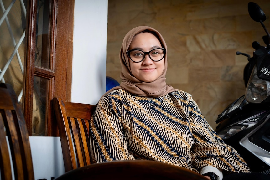 A close up of a smiling woman sitting in a chair while wearing a hijab and glasses.