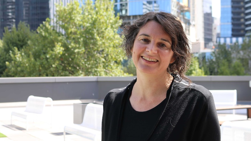 A woman with short brown hair and black shirt and jacket looks to camera smiling broadly, with blurred cityscape in background.