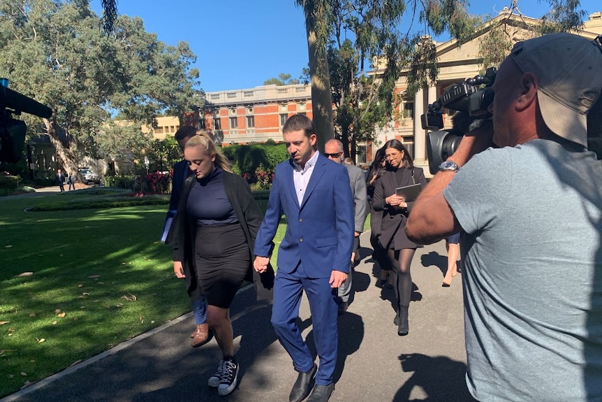 Kyle Hartigan holds hands with a blonde lady as he is flanked by cameras leaving court