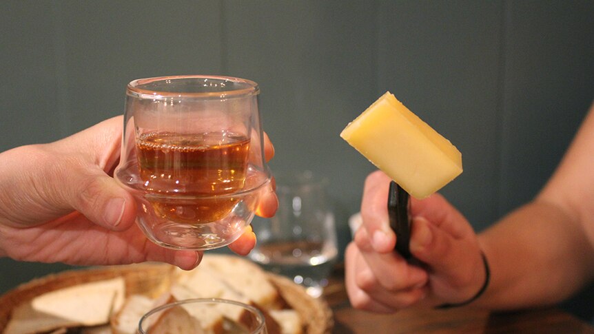 A hand holding clear glass mug full of amber-coloured tea beside a hand holding a wedge of hard cheese on a black-handled knife.