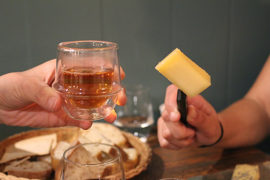 A hand holding clear glass mug full of amber-coloured tea beside a hand holding a wedge of hard cheese on a black-handled knife.