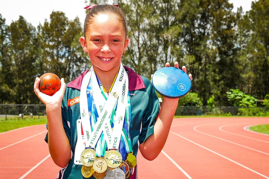 A young girl wearing many medals holds up a shot put and discus while standing on a running track