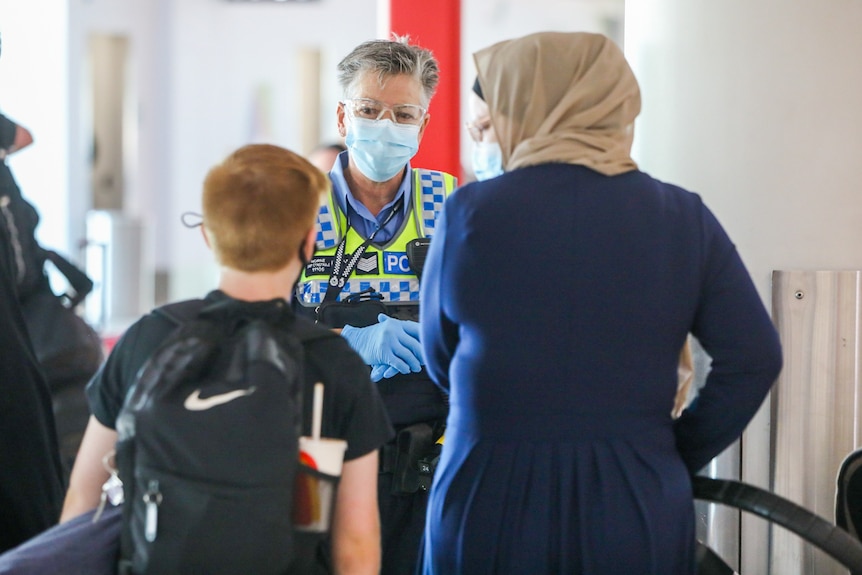 A police officer wearing a mask speaks to two people with their backs to the camera.