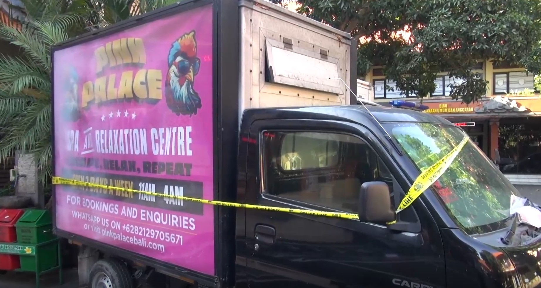 A pink and black truck with a bright advertisement for the Pink Palace sits outside a building with yellow tape around it.