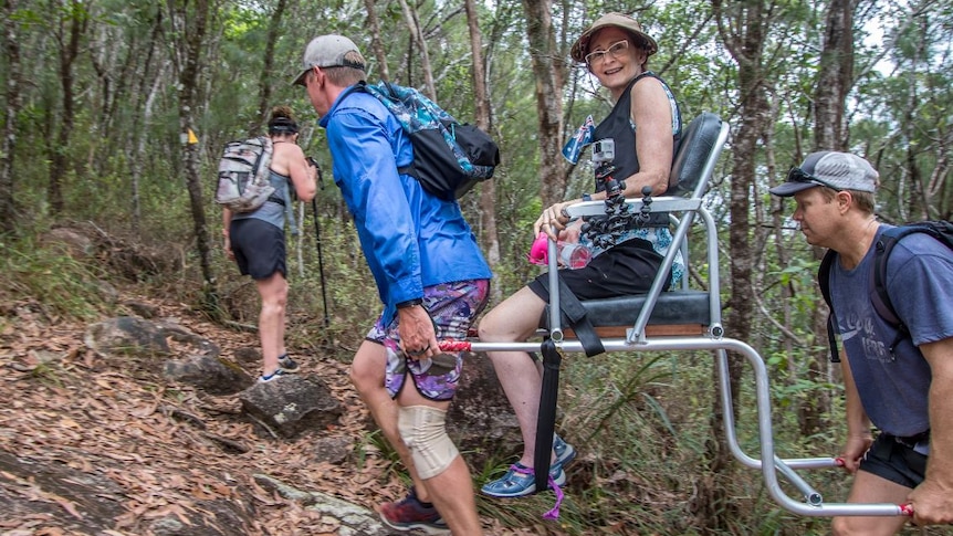 Two men carry a woman sitting in a specially-built chair up a bush track.