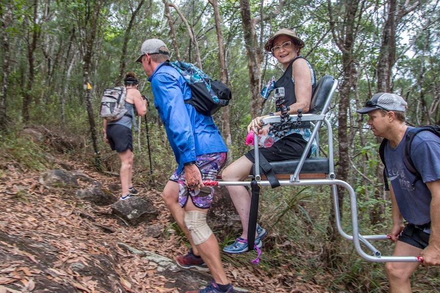 Two men carry a woman sitting in a specially-built chair up a bush track.