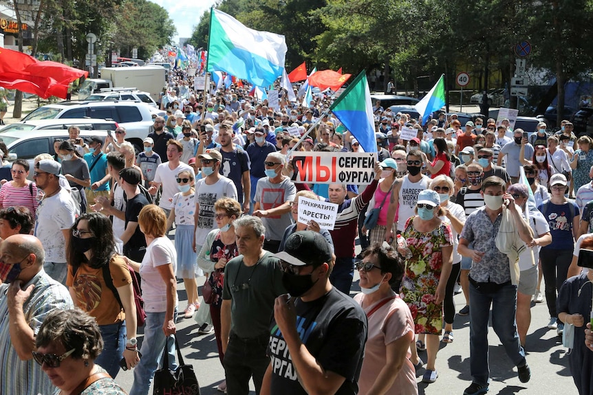 A large crowd of people wave flags and signs while rallying along a road in the sunshine.