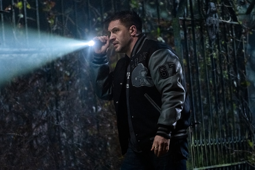 Man with dark hair in black and grey bomber jacket walks towards something menacing outside with a flashlight in his hand.