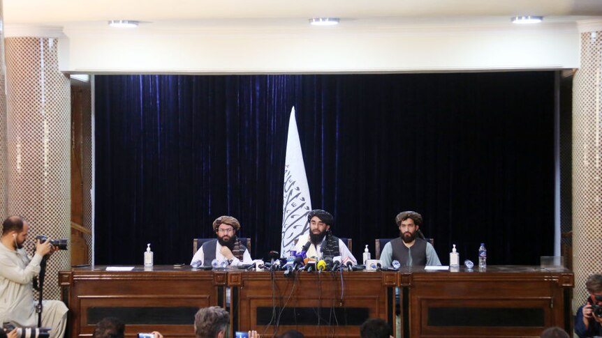 three men sit in front of many microphones at long table with taliban flag in background surrounded by the press