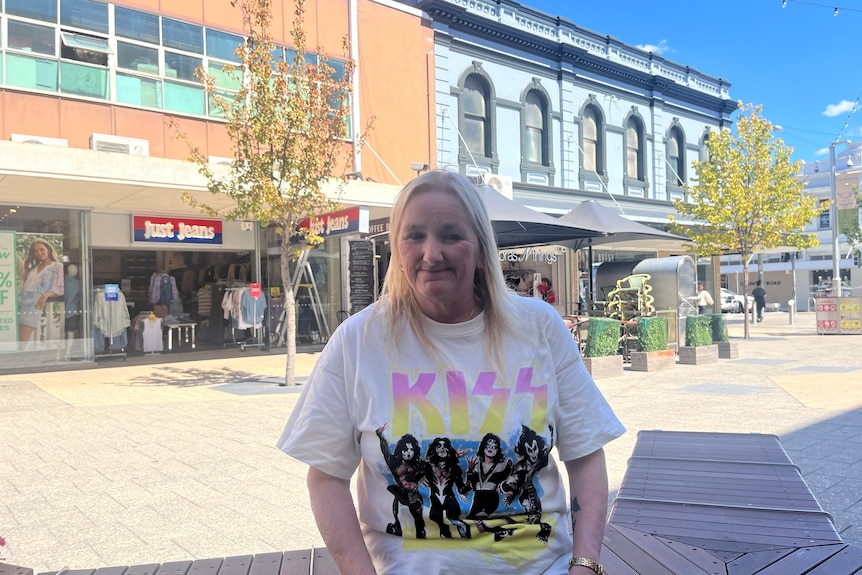 A woman with long blonde hair wearing a Kiss t-shirt sits on a wooden seat in a shopping mall.