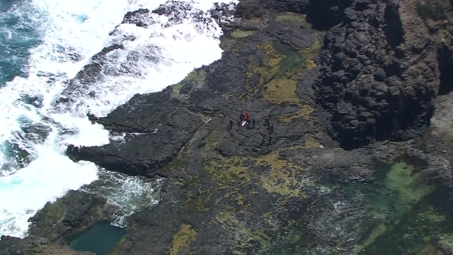 Una vista aérea desde la distancia de personas reunidas alrededor de una camilla sobre rocas mientras las olas del océano rompen debajo.