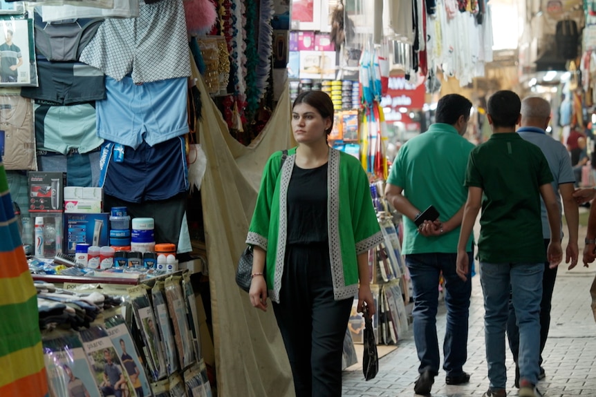 a woman in a green jacket walks through an indoor market past stalls selling clothes