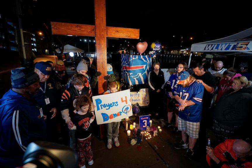 NFL fans in Cincinnati Bengals and Buffalo Bills gear pray next to a cross in front of a hospital with a 'Prayers 4 Damar' sign.