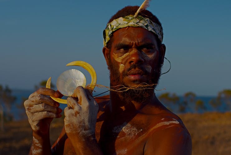 An Aboriginal man in traditional dress holds a carved shell up to the light