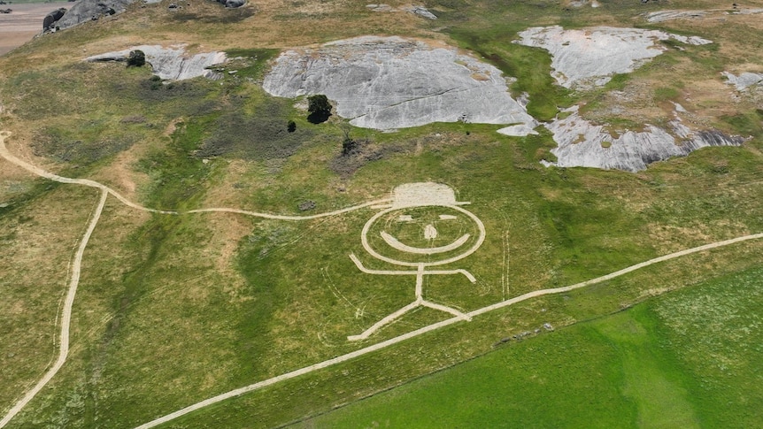 Aerial shot of large stick figure, smiley face, mowed into the side of a green hill.