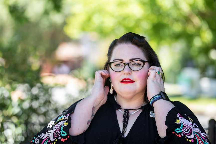 A woman with black hair, black glasses and red lipstick stands outside with her fingers in her ears.