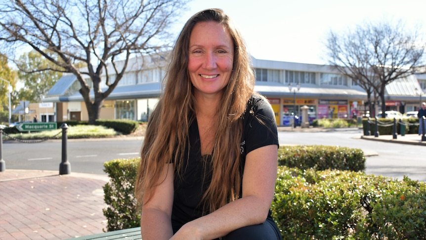 A lady with long brown hair smiles at the camera. Behind her is a regional city street.