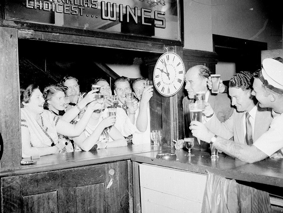 A 1950s black-and-white photo of excited pub patrons raising their glasses as a clock nears 10pm