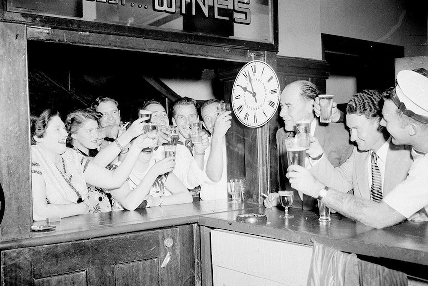 A 1950s black-and-white photo of excited pub patrons raising their glasses as a clock nears 10pm
